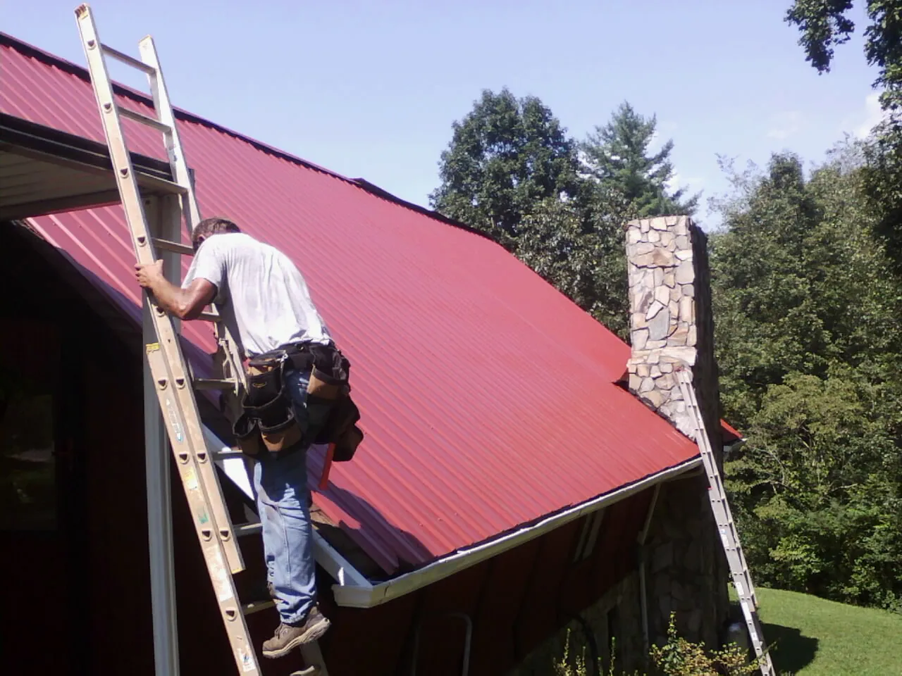 Roofer on a ladder beside a red metal roof and stone chimney — Soffit & Fascia Repair by Roofing Brothers in Odessa